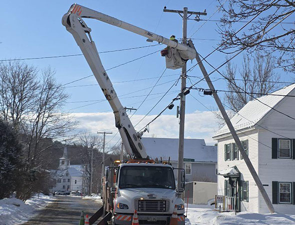 A utility truck with a raised bucket lift repairs power lines on a snowy street, surrounded by houses and trees.