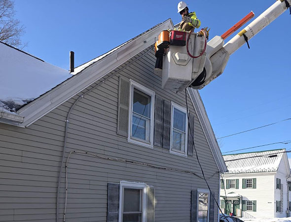 A worker in a bucket lift clears snow from the roof of a beige house on a snowy day, with blue skies in the background.