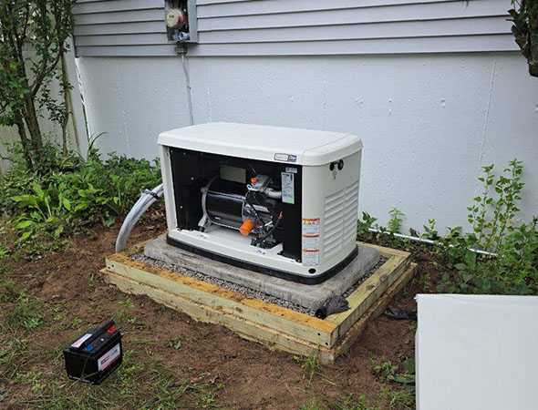 A generator on a wooden platform, partially open, with a battery nearby, surrounded by grass and vegetation against a house wall.